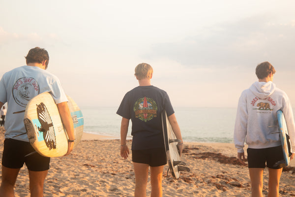 A photo of three Joe's Surf Shop surfers heading towards the waves. Each are wearing their own Joe's Surf Shop apparel; the surfer on the right is wearing the Joe's Surf Shop Papa Joe Tee; middle is wearing the Joe's Surf Shop Longboard Tee; and the right is wearing the Joe's Surf Shop Surfing Bear Hoodie.